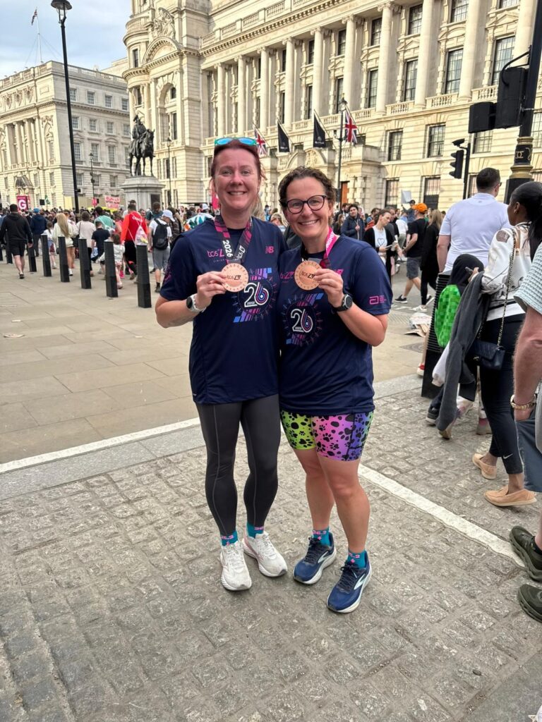 Two smiling runners pose together on a city street, each holding a medal. They wear matching navy race shirts and sports gear, with a historic building and a crowd of people in the background.