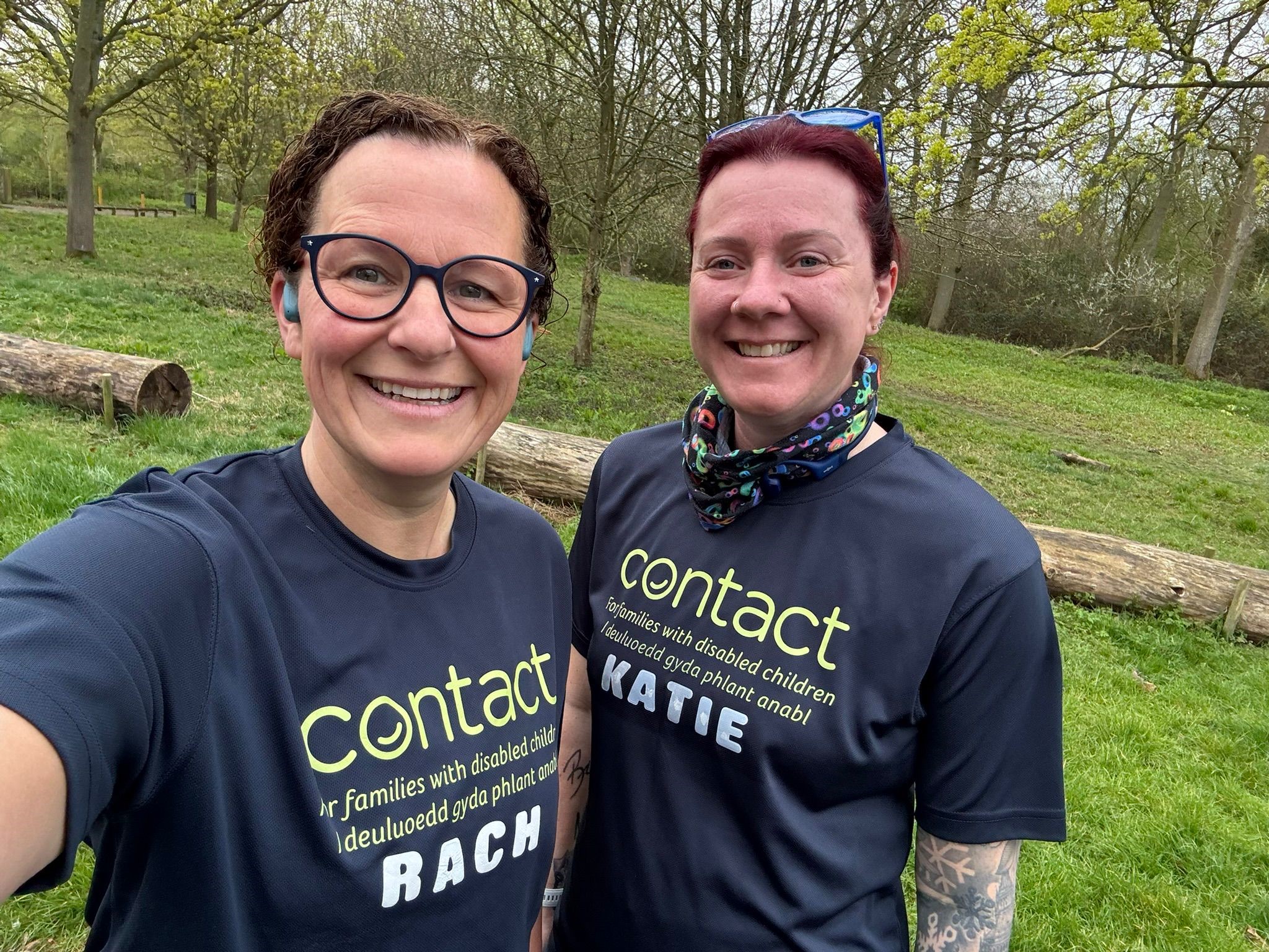 Two people smiling outdoors on grass, wearing navy "Contact" charity shirts. One shirt says "RACH" and the other "KATIE." There are trees and fallen logs in the background on a cloudy day.