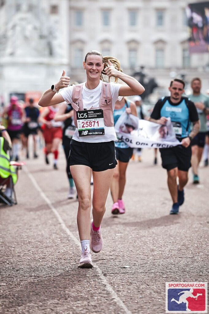 A smiling runner gives two thumbs up whilst running in a race, wearing a pink shirt and black shorts. Other runners and a large white monument are visible in the background.