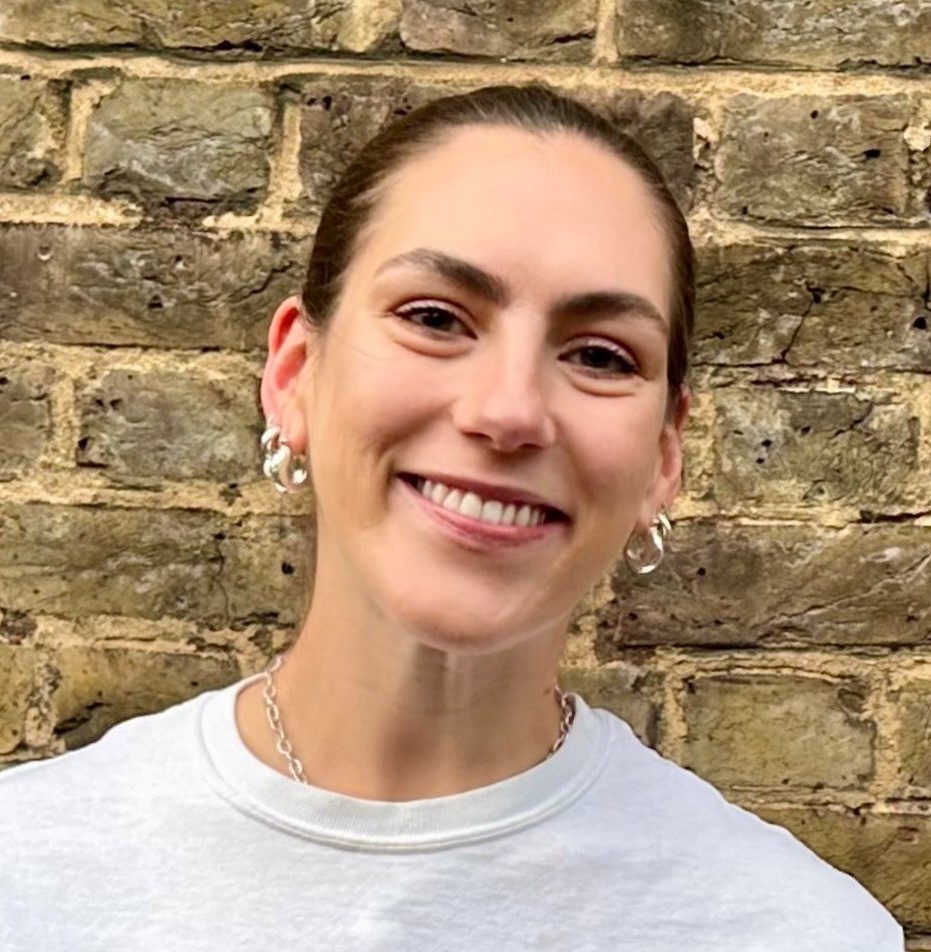 A woman with brown hair tied back smiles at the camera, standing in front of a weathered brick wall. She is wearing a white shirt, silver hoop earrings, and a chain necklace.