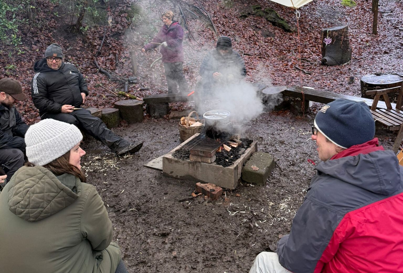 A group of people sit on logs around a smoky campfire in a muddy, wooded area. Food is cooking on the barbeque, and the setting looks wet and chilly. Some people wear hats and jackets.