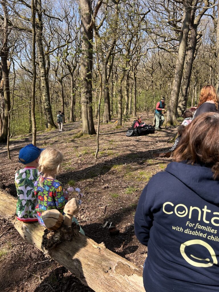 Children and adults sit on a log and stand in a free woodland area, watching a person with outdoor kit. One adult wears a hoodie that says "contact, For families with disabled children." Sunlight filters through the trees.
