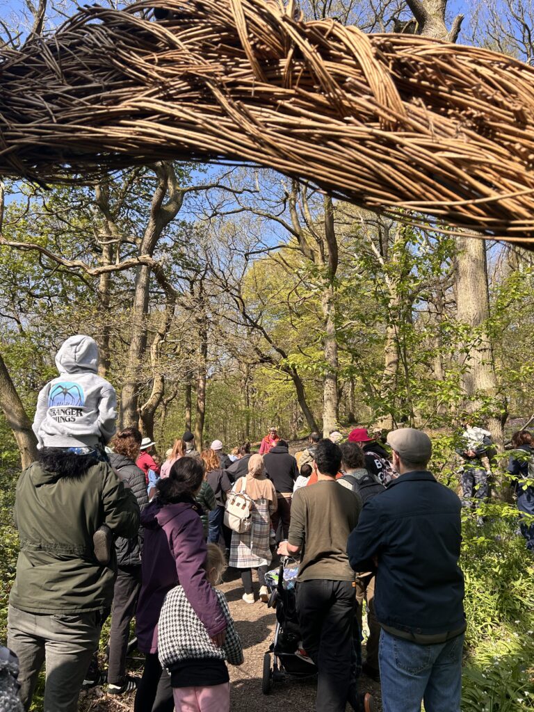 A crowd of people, some with children, walk along a free woodland path under a large woven arch made of branches. Sunlight filters through the trees, and many are wearing jackets, suggesting a cool day.