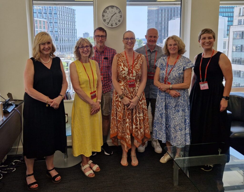 Seven adults stand together smiling in an office with large windows showing city buildings. A wall clock above reads 12:15. The group, discussing child trust fund providers, wears summer clothing and conference lanyards for a professional vibe.