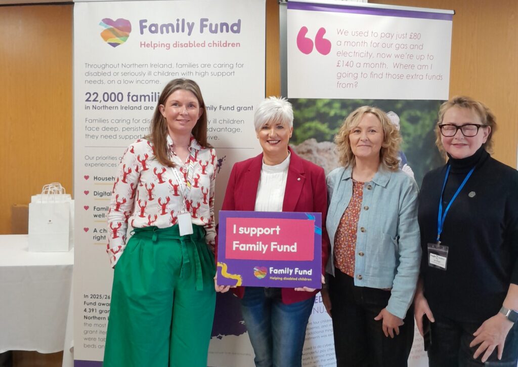 Four women stand smiling at an event in front of Family Fund banners. One woman holds a sign that reads "I support Family Fund." The banners highlight support for families with disabled children.