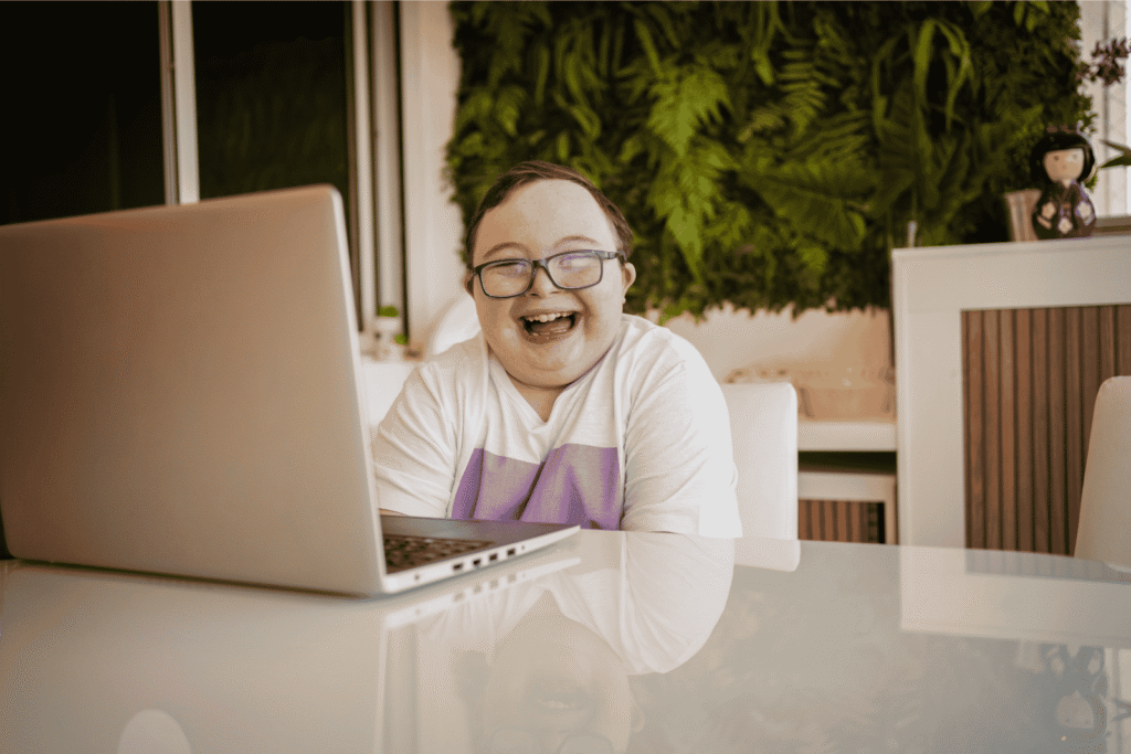 A person with Down’s syndrome sits at a table using a laptop, smiling joyfully. There is greenery on the wall behind them and a small decorative doll on a cupboard.