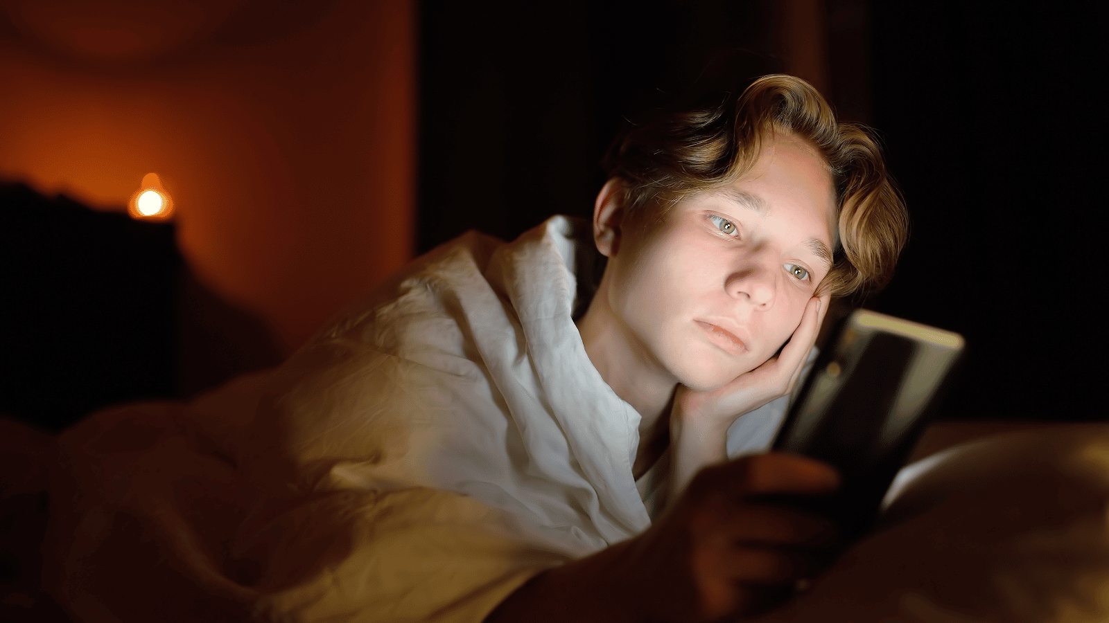 A person with light brown hair lies in bed under a white duvet, resting their head on one hand while looking at a mobile phone screen. The room is dimly lit with a soft, warm glow in the background.