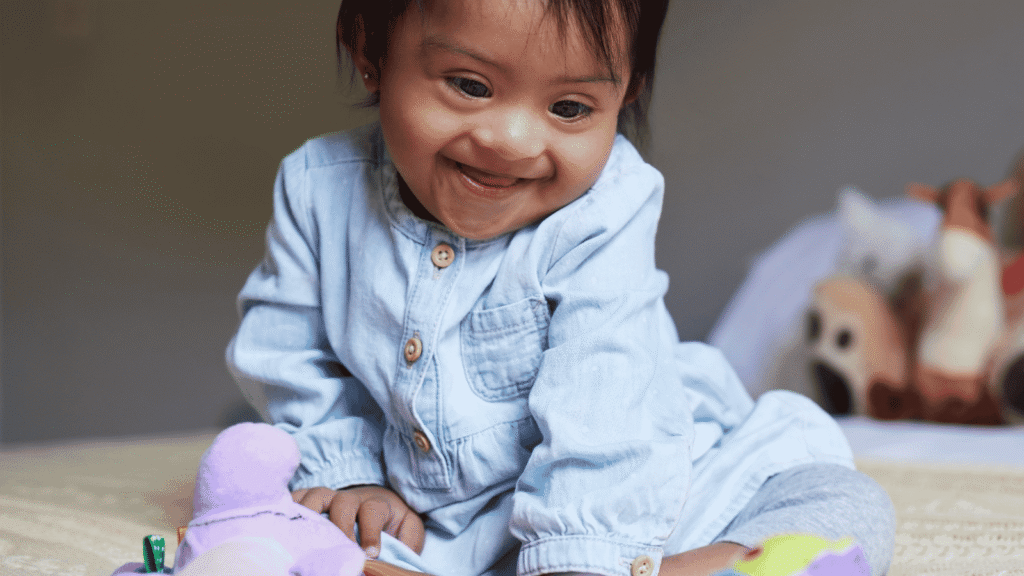 A smiling toddler wearing a light blue dress sits on a bed, looking down at colourful soft toys in front of her. The background is softly blurred.