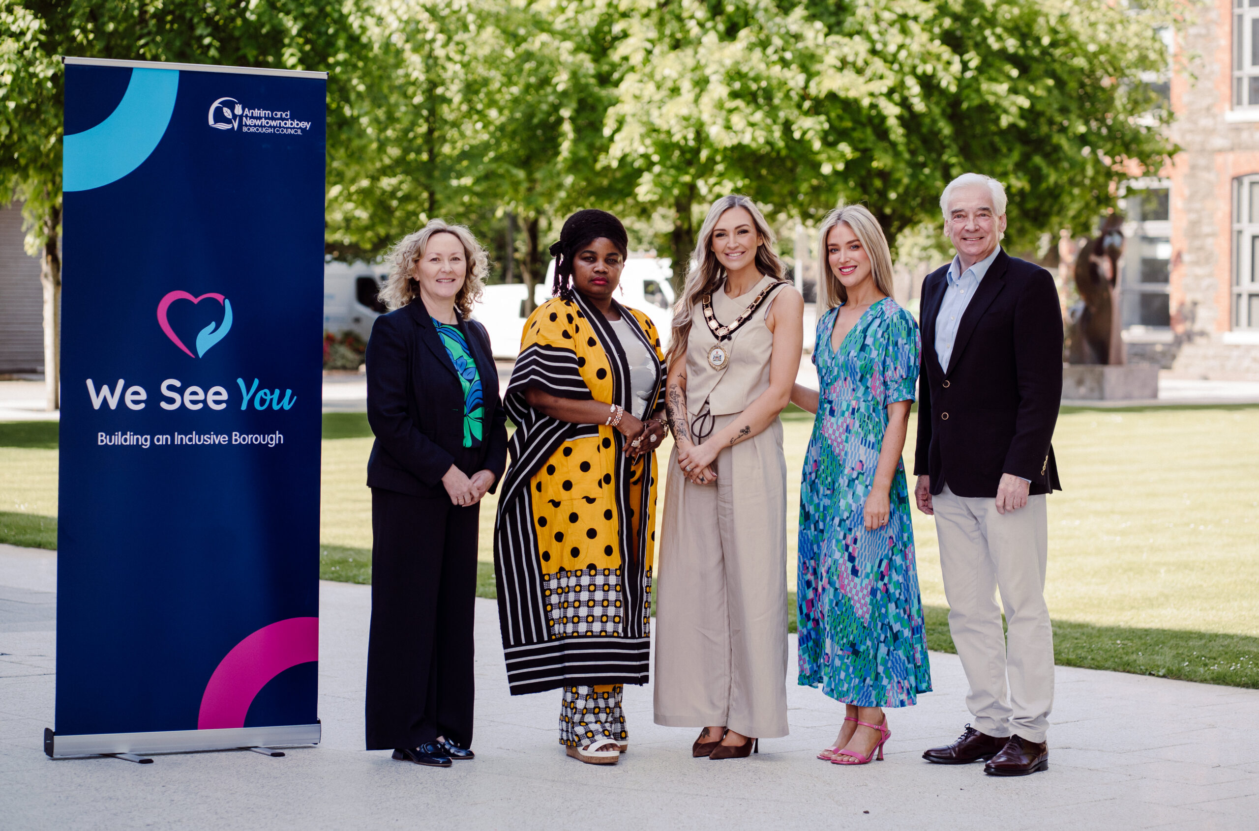 Five people stand outdoors beside a sign that reads "We See You: Building an Inclusive Borough." They are dressed in smart attire, smiling, with trees and a building in the background on a sunny day.