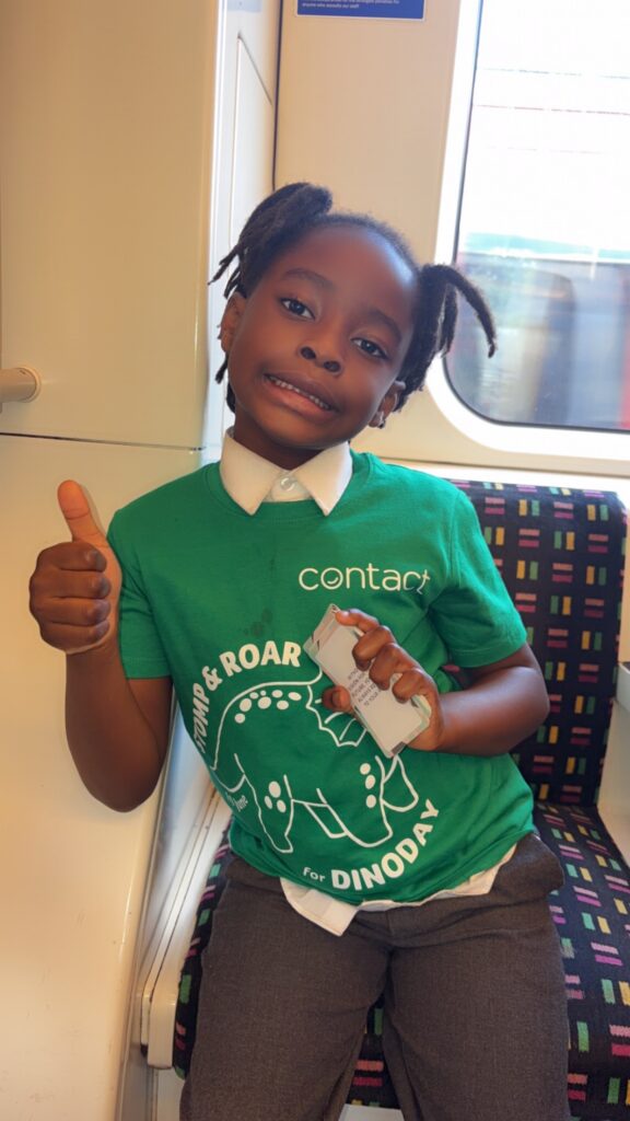 A young child sits on a train seat, smiling and giving a thumbs up. The child is wearing a green "Contact" t-shirt with a dinosaur graphic and is holding a white object in their other hand.