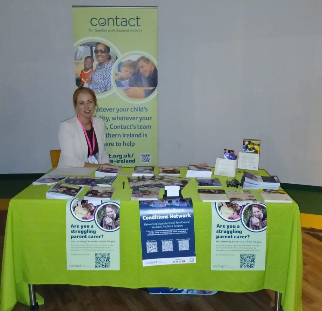 A woman sits behind a table covered with leaflets and posters about support for families with disabled children, under a 