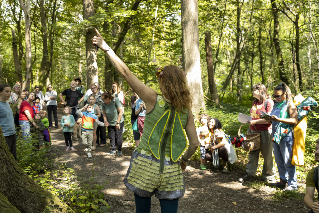 A woman dressed in green fairy wings gestures upwards while a group of adults and children watch her in a sunlit forest. Some people are holding papers, and others appear to be listening or engaging with the scene.