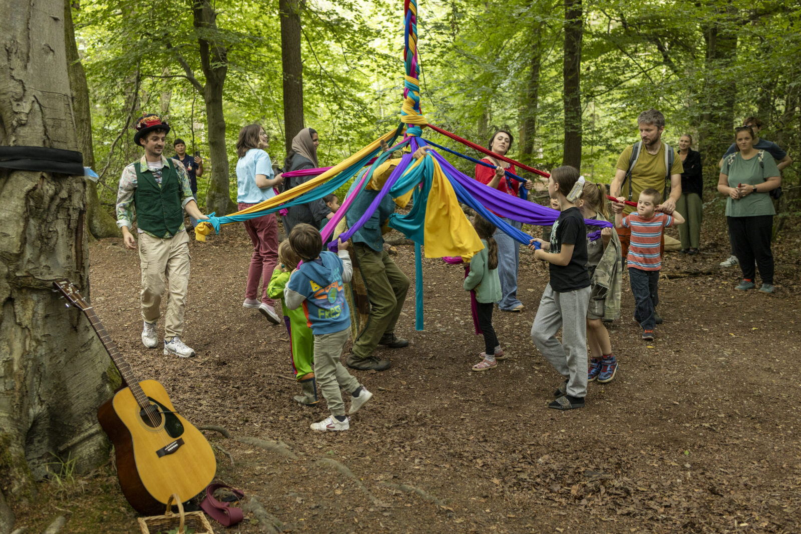 A group of children and adults dance around a maypole in a forest, holding colourful ribbons. A guitar rests against a tree nearby. The scene is lively and joyful among the green trees.