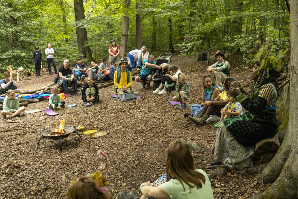 A group of people sit in a forest circle around a small fire pit, listening to a storyteller. Adults and children are present, surrounded by trees and greenery, creating a communal, peaceful atmosphere.