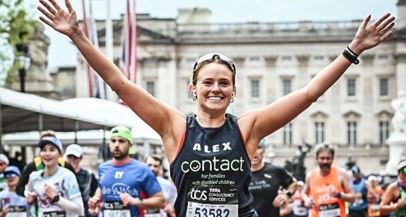 A smiling runner with arms raised crosses a marathon finish line, wearing a bib and black vest reading 