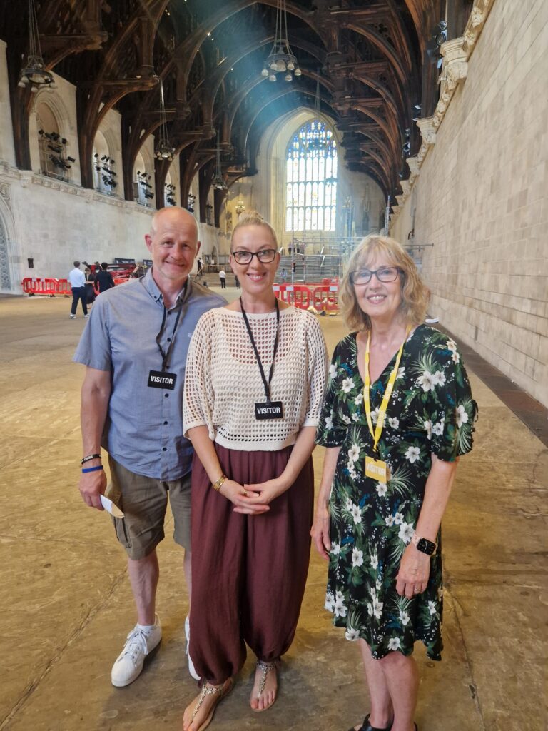 Three adults wearing visitor badges stand smiling inside a large, historic hall with arched wooden beams, stone walls, and a big stained-glass window in the background.