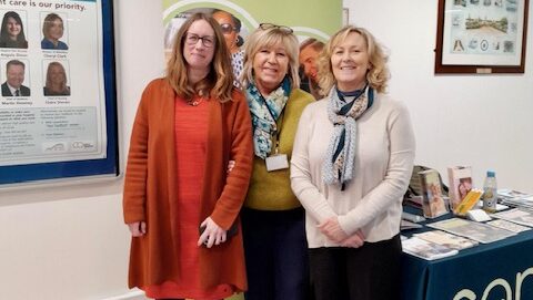Three women stand indoors, smiling at the camera. Behind them is a display board with photos and brochures on a table. They are dressed in casual, professional attire and appear to be at an event or office setting.
