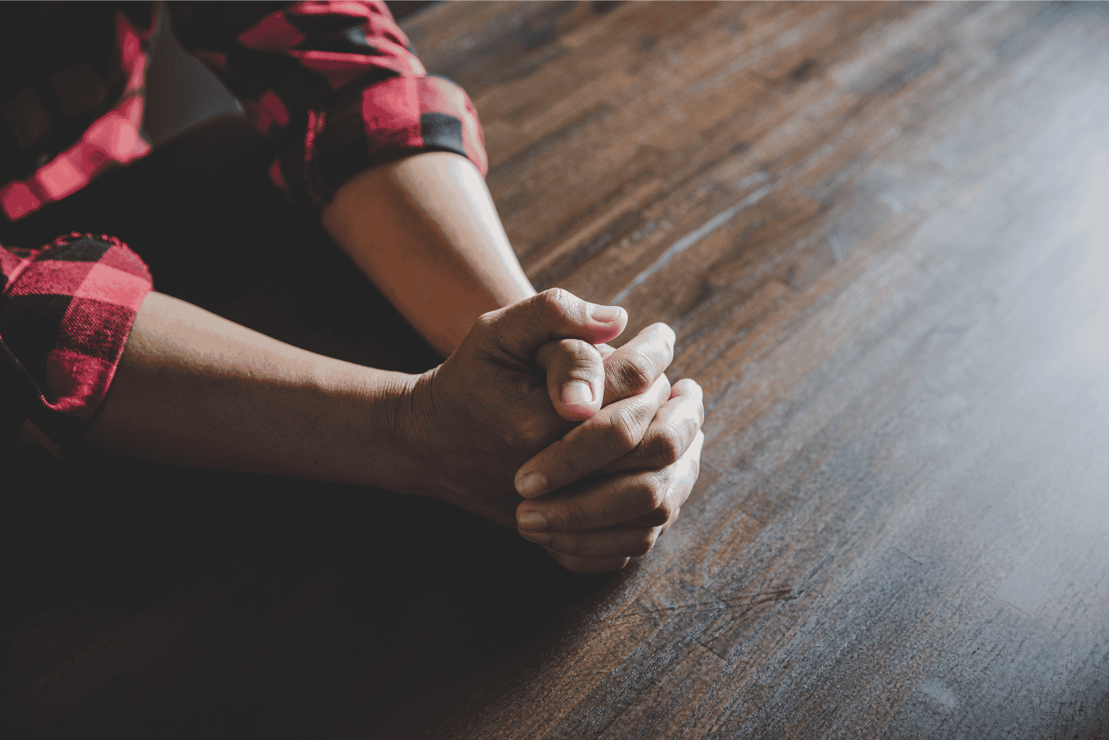 A person in a red checked shirt sits at a wooden table with their hands clasped together, resting on the surface.