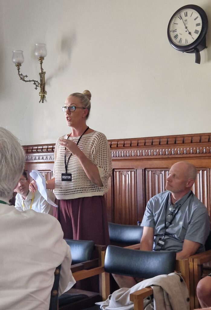 A woman wearing glasses and a "VISITOR" badge stands and speaks during a child trust fund round-table in a wood-panelled room with other seated people. A clock and wall lamps are visible on the wall behind her.