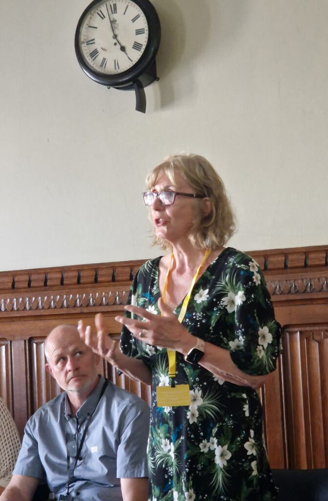 A woman in glasses and a floral dress speaks, gesturing with her hands, while a man sits and listens beside her during a child trust fund roundtable.