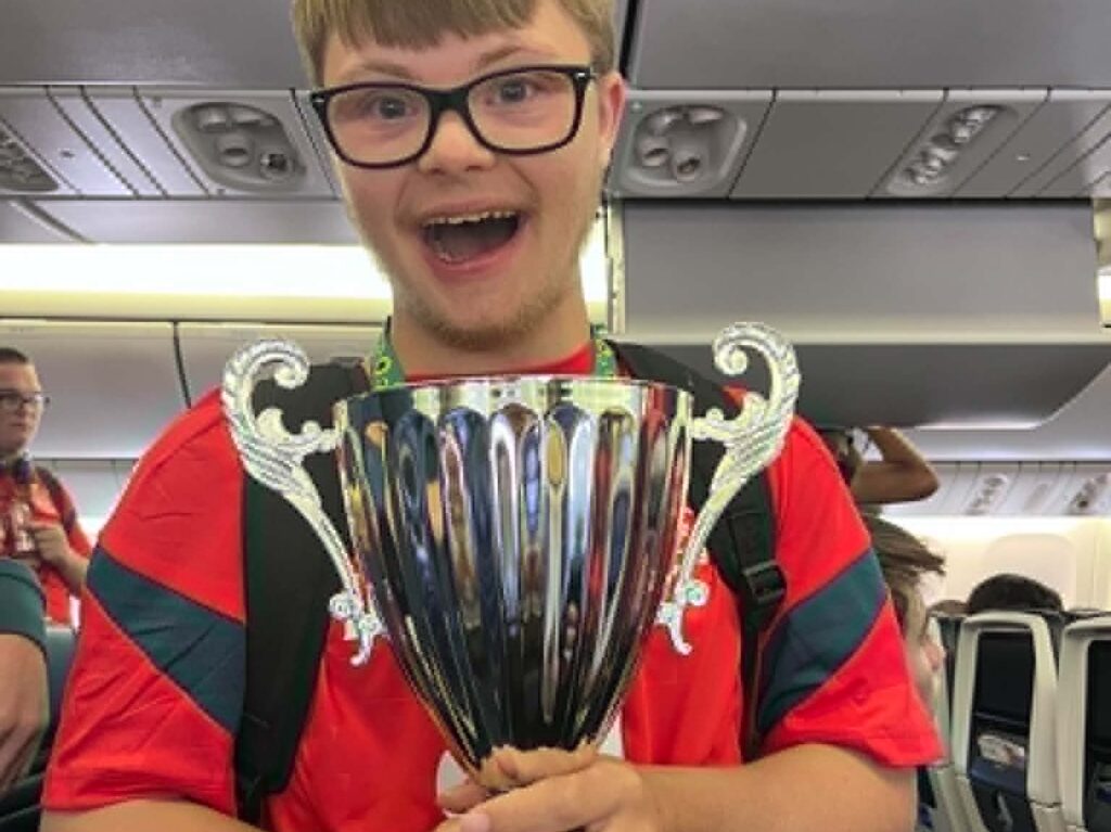 A smiling young person with glasses and a red shirt holds a large trophy on an aeroplane, with other passengers visible in the background.