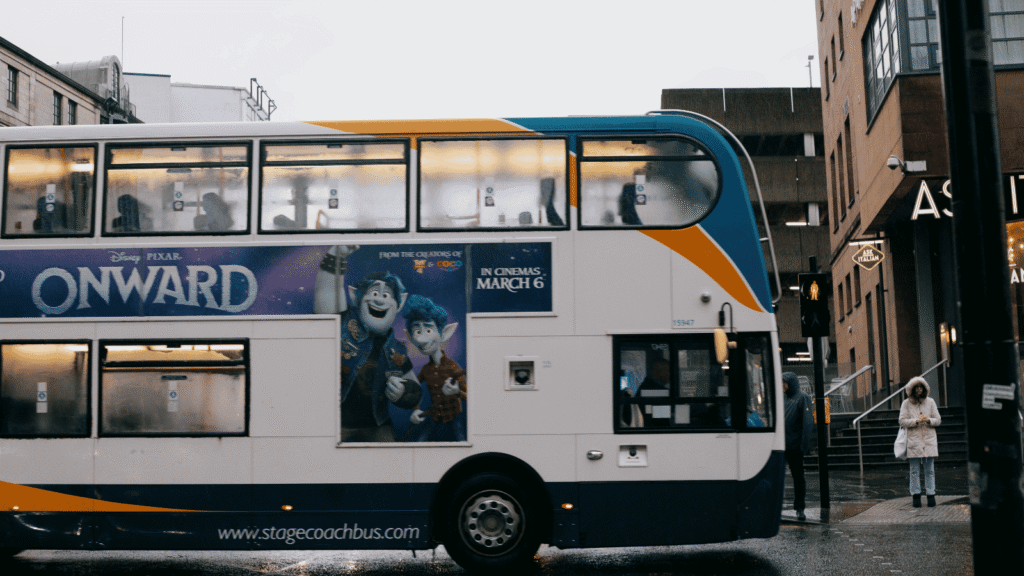 A double-decker bus with an “Onward” film advert is stopped on a rainy city street. People are visible through the bus windows; a person in a white coat stands on the pavement holding an umbrella.