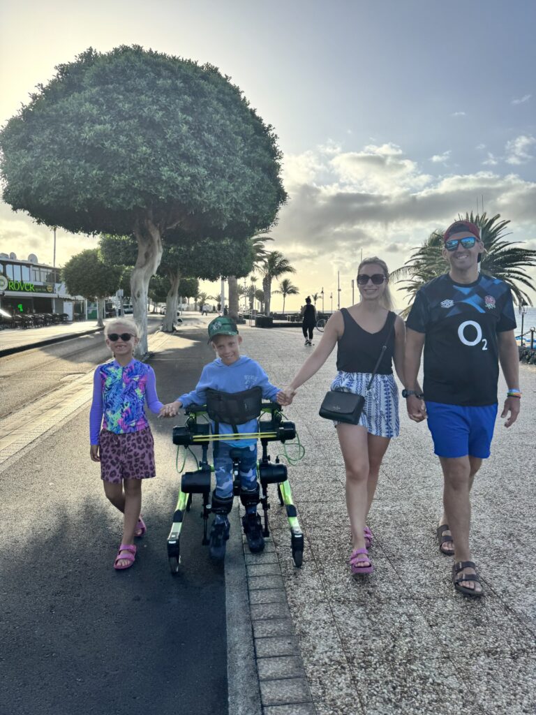 A family of four walks outside on a sunny day. A young boy uses a green walking frame, holding hands with his sister and parents. Palm trees and a large leafy tree line the path.