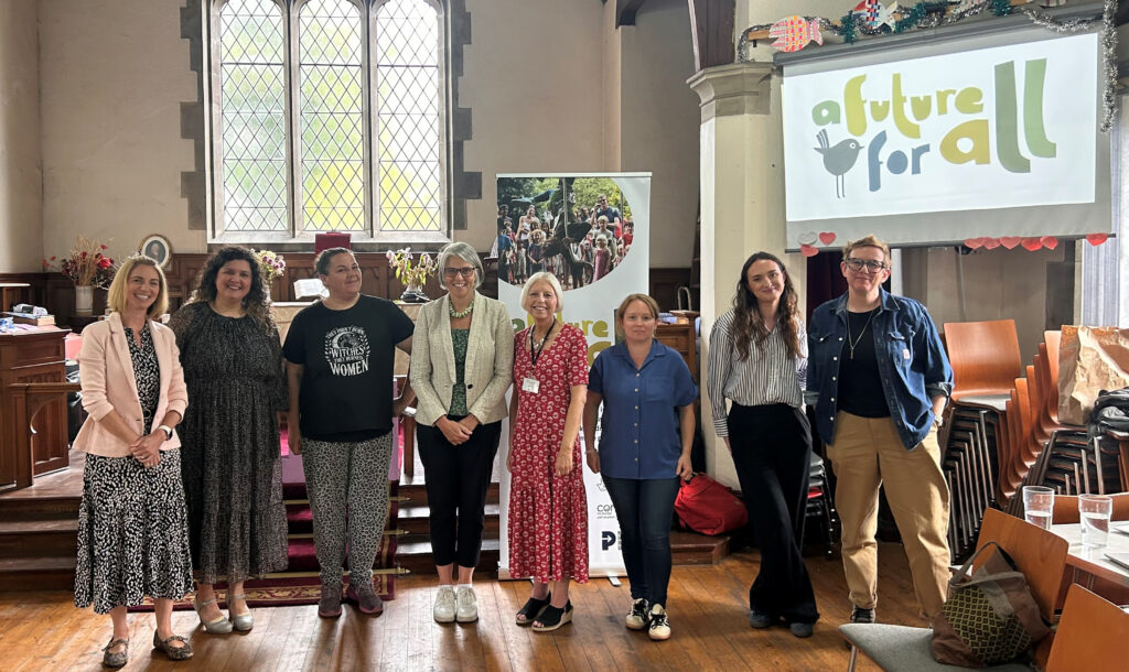 Eight people stand side by side in a hall with a large window, wooden floor, and an “A Future for All” banner behind them. They are smiling and dressed in a variety of outfits.