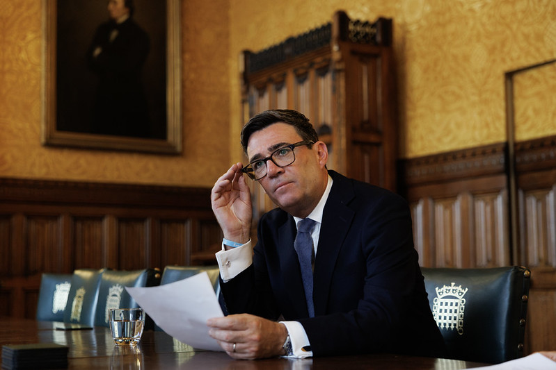 Prime Minister Andy Burnham in a suit and glasses sits at a boardroom table holding a document as the Prime Minister addresses a child trust fund.