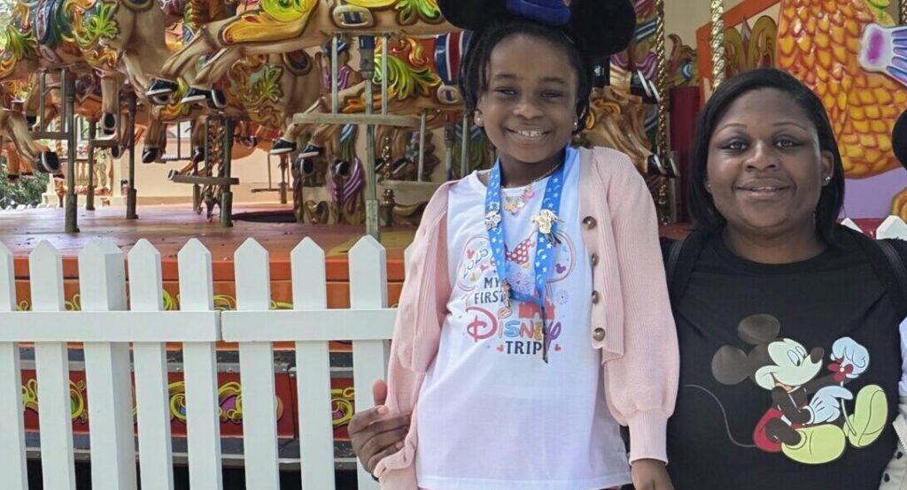 A young girl wearing Mickey ears and a “My First Disney Trip” shirt standing with a woman in front of a roundabout.